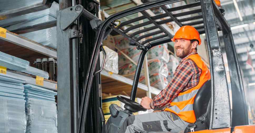 Funcionário de capacete laranja foca no uso de EPIs na empilhadeira para prevenir riscos.