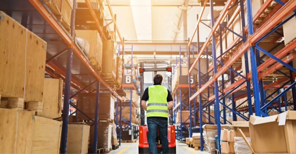 Operador de colete guia empilhadeira em galpão de logística com supply chain.